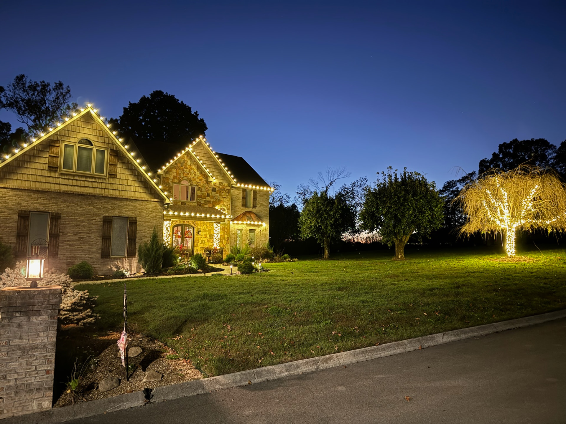 Two story brick and stone home with warm white C9 roofline lighting and a lit weeping willow tree seen from the street at dusk