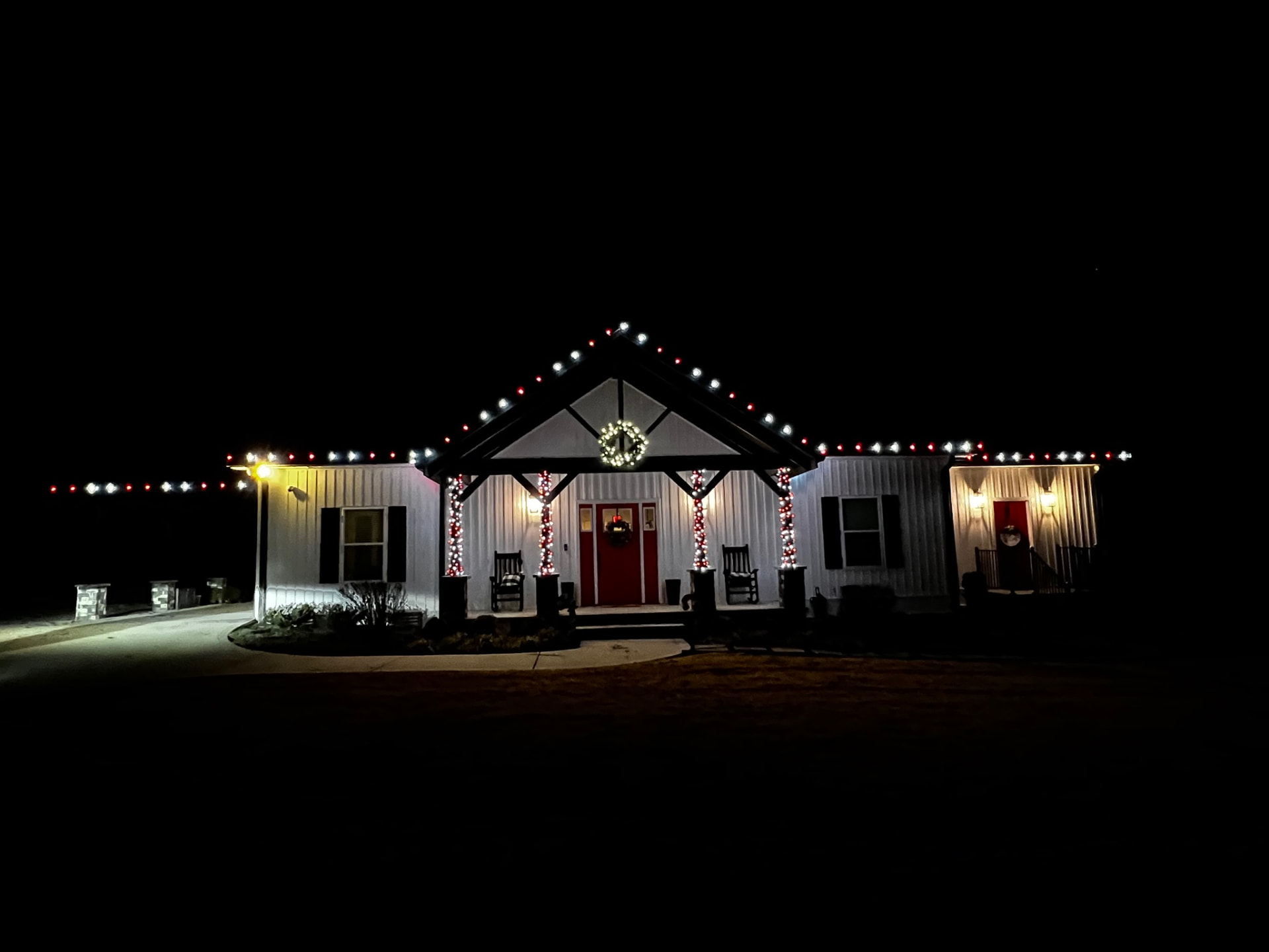 White metal barndominium home at night with red and white C9 Christmas lights on the roofline, candy cane wrapped porch posts and a lit wreath