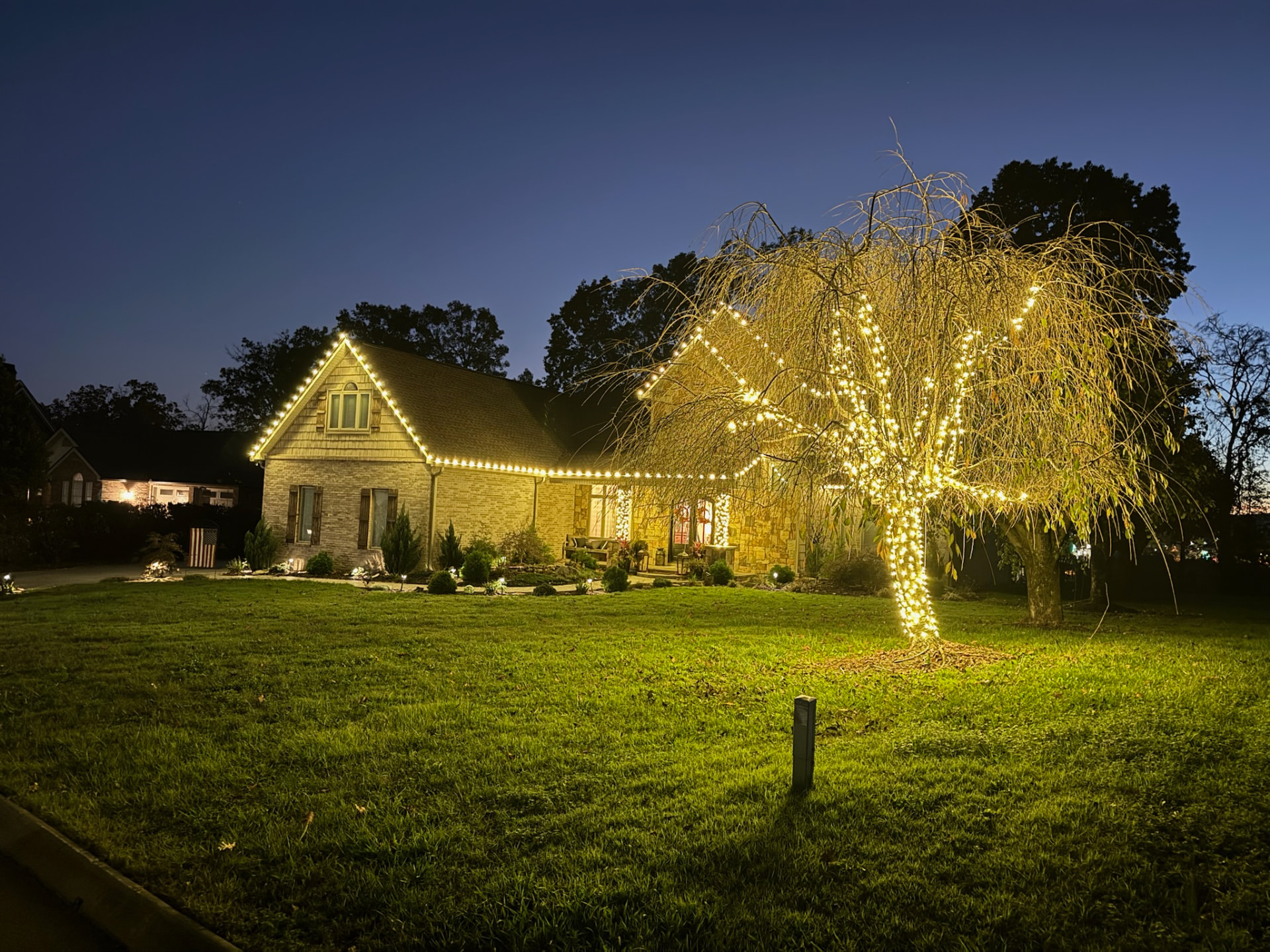 Weeping willow tree fully wrapped in warm gold lights beside a brick Maryville TN home outlined in warm white C9 Christmas lights at dusk