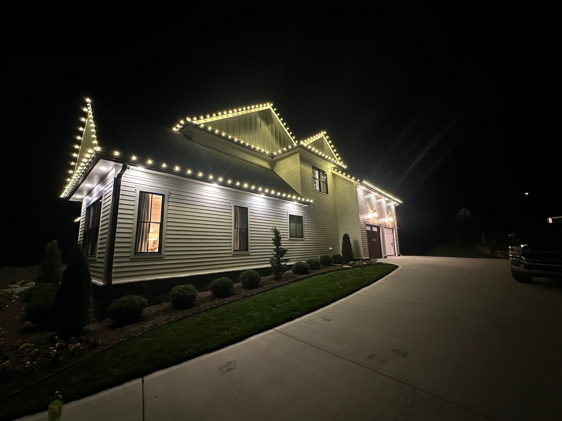 Side elevation of a farmhouse style home lit with warm white C9 Christmas lights along dormers, eaves and the garage roofline