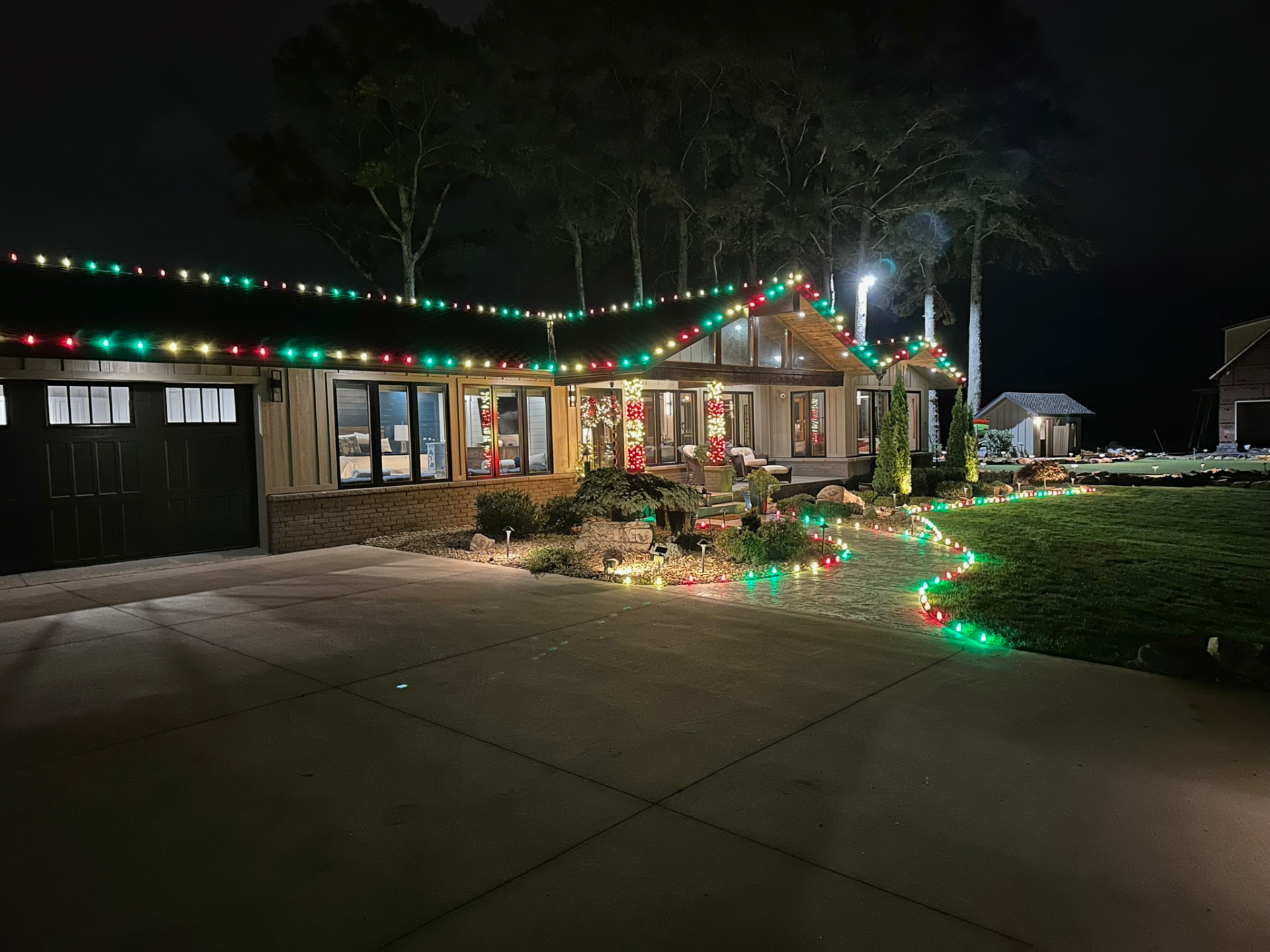 Modern ranch home at night with red green and white C9 roofline lights, candy cane wrapped porch columns and a lit curving walkway