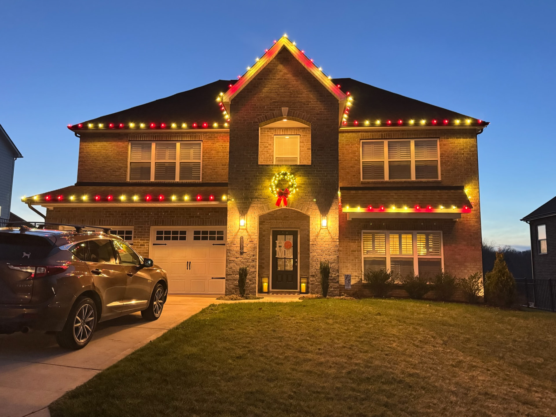 Two story brick home at dusk with alternating red and warm white C9 Christmas lights on every roofline plus a lit wreath over the entry